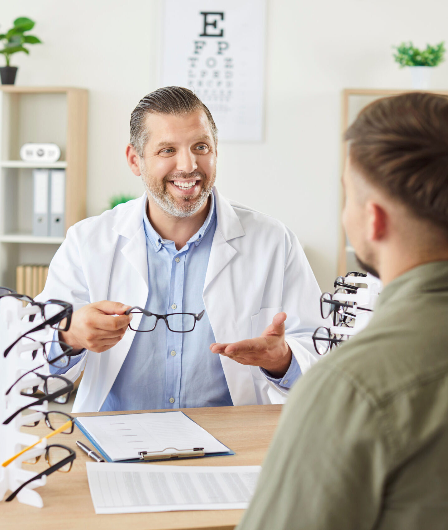 Male mature ophthalmologist doctor consulting patient in ophthalmology clinic holding spectacles in hands in office. Young man choosing eyeglasses in optic store. Eye health concept.