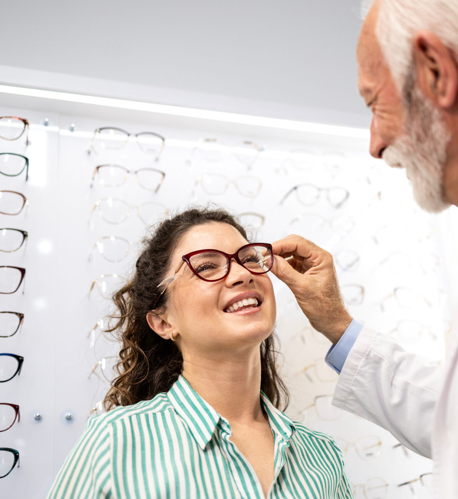 Beautiful smiling brunette woman trying on fashionable eyeglasses in optician store with help of experienced senior optometrist.